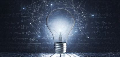 A lightbulb illuminated in front of a dark background with bits of light floating around the bulb