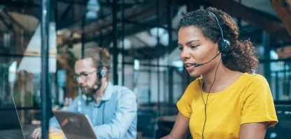 Two support agents reviewing information on their computers while wearing headsets in a modern office building room